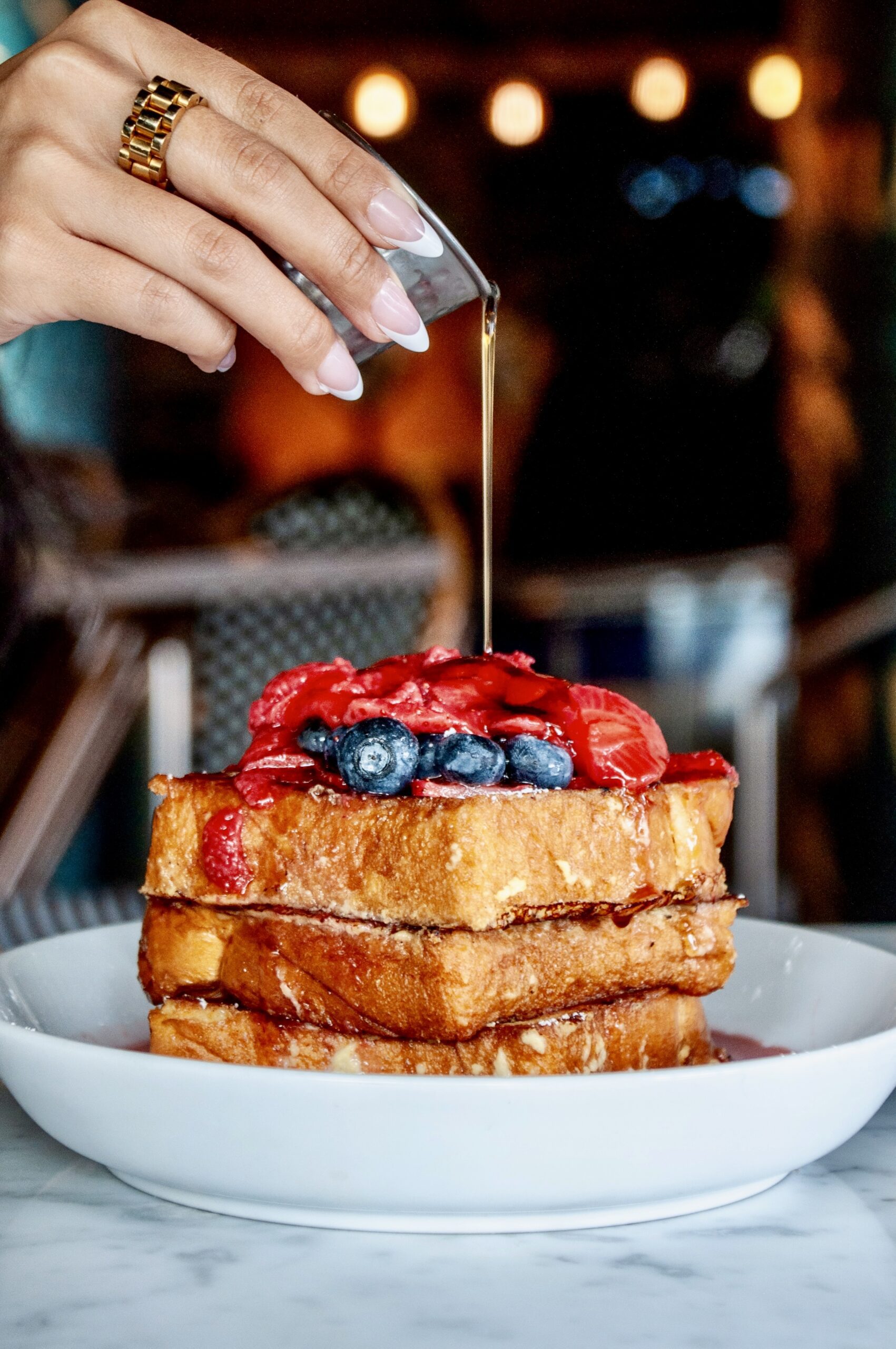 Hand pouring syrup on golden brown French toast topped with fresh berries and powdered sugar at Puttery.